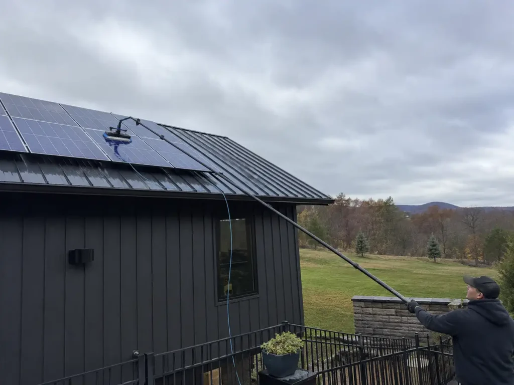 A professional cleaner using a long carbon fiber telescoping pole with a brush head to clean rooftop solar panels